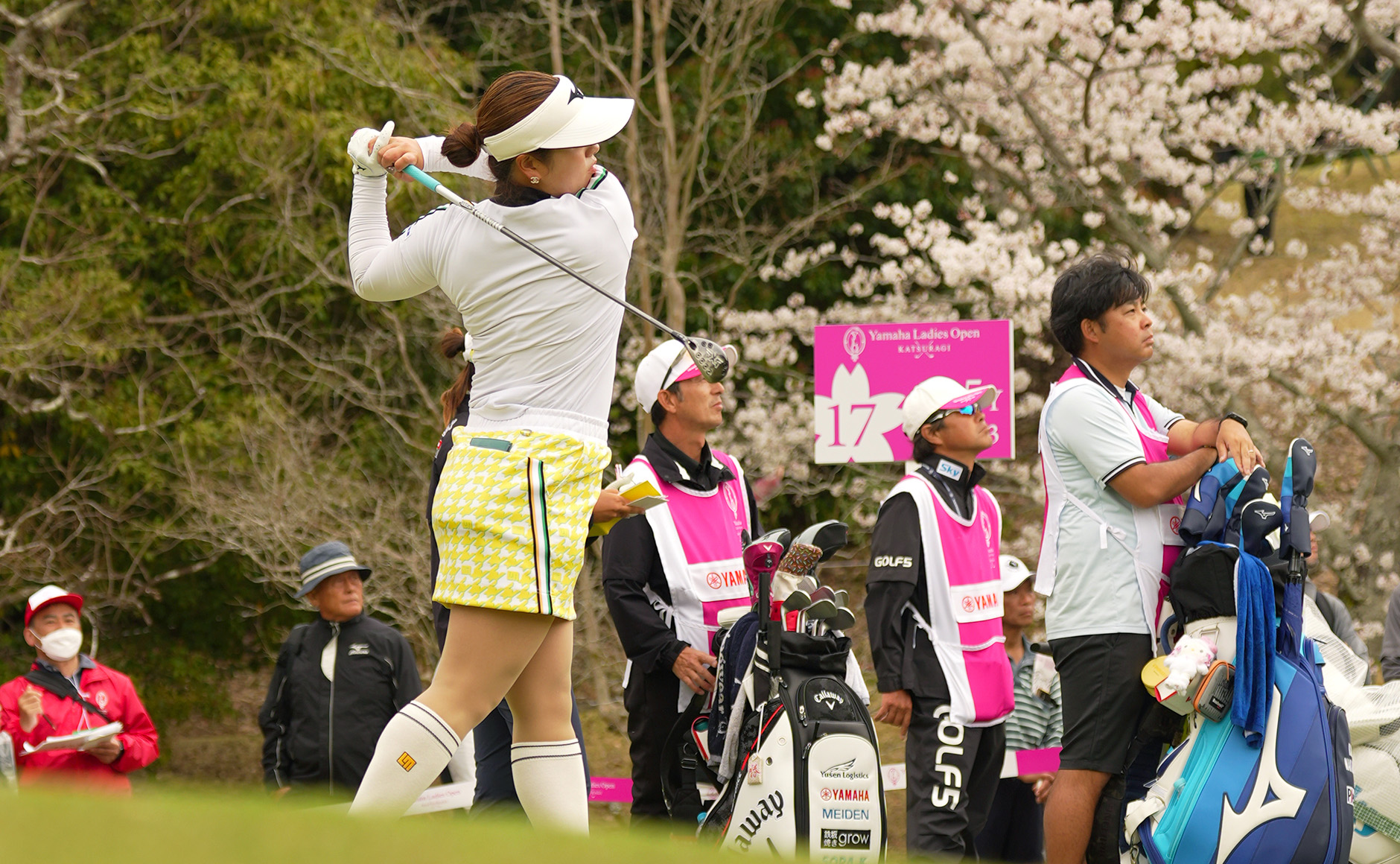 満開の桜を背にショットを打つ仲村果乃と、ボールの行方を追うキャディやギャラリー